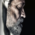 Primer plano lateral del rostro de un hombre barbado, iluminado con fuerte contraste sobre fondo oscuro.