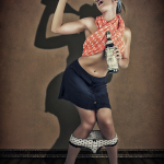 Mujer adulta con pañuelo naranja al cuello y falda azul, posando de pie con gesto pin-up sobre fondo marrón.