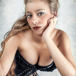 Mujer adulta rubia con corsé negro, apoyando el rostro en la mano en un retrato claro de estilo pin-up.