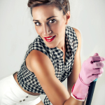 Mujer adulta con blusa de lunares y guantes rosas, inclinada hacia la cámara en una escena doméstica de estética pin-up.