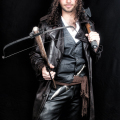 Hombre adulto vestido de negro, sosteniendo varias armas o espadas en una pose de cazador gótico.