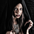 Mujer adulta con capucha negra y mirada intensa, en una escena gótica de penumbra.