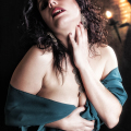 Mujer adulta con vestido verde azulado caído sobre el hombro, en una escena sensual y oscura.