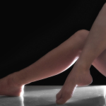 Piernas desnudas apoyadas en el suelo con un pie adelantado, sobre fondo negro y luz lateral.