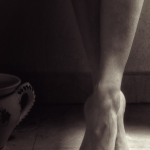 Pie desnudo apoyado en puntas junto a una pared, con luz suave y fondo oscuro.
