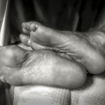 Composición en blanco y negro de dos pies desnudos cruzados o entrelazados sobre una cama.