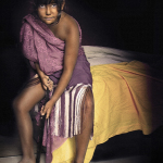 Niña sentada sobre una cama, envuelta parcialmente en una tela malva, en una escena íntima de inspiración pictórica.