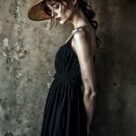 Mujer adulta con vestido negro y sombrero de ala ancha, posando de perfil ante una pared envejecida.