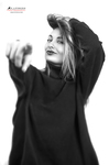 Retrato en blanco y negro de mujer adulta con sudadera oscura y capucha, levantando una mano en una pose expresiva.