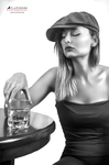 Retrato en blanco y negro de mujer adulta con gorra y vaso, apoyada en una mesa con una botella a su lado.