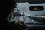 Hombre conduciendo un coche bajo la lluvia, visto desde el interior del vehículo, con la carretera mojada y el mar al fondo.