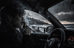 Hombre fumando en el interior de un coche, con la ventanilla cubierta de lluvia y el mar oscuro al otro lado.
