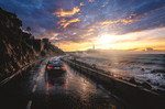 Coche negro avanzando por una carretera costera mojada al atardecer, con el cielo iluminado y el faro en la distancia.