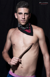 Gustavo posa sin camiseta con pañuelo al cuello y vara en la mano, sobre fondo negro de estudio.