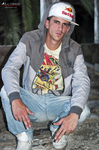 Gustavo sentado con ropa informal, gorra blanca y chaqueta gris, en una escena urbana relajada.