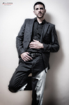 Gustavo posa con traje negro apoyado sobre fondo claro, en una imagen de moda masculina.