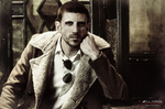 Retrato de Gustavo con chaqueta clara y gafas de sol colgadas, en una escena de tonos sepia.