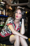 Esther sentada con las piernas cruzadas en un bar, con blusa estampada y expresión sugerente.