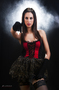 Modelo femenina sentada con corsé rojo y falda negra, en una fotografía de estudio con fondo oscuro.