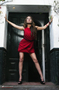 Modelo femenina con vestido rojo apoyada en una puerta, en una escena urbana.