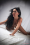 Modelo femenina morena tumbada en una cama blanca, mirando a cámara.