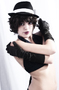 Modelo femenina con sombrero negro y ropa oscura, en una pose de estudio.