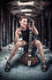 Modelo femenina sentada con guitarra en un entorno oscuro de ladrillo.