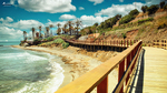 Sendero Litoral de Mijas junto al mar, con pasarela de madera, playa, palmeras y edificios costeros al fondo.