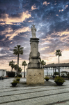 Monumento en una plaza de Rota, Cádiz, con palmeras, farolas y cielo dramático al fondo.
