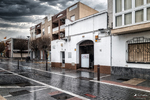 Calle de Rota con fachadas blancas, comercios y suelo mojado bajo un cielo gris.