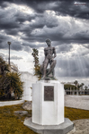 Escultura sobre pedestal blanco en Rota, Cádiz, con paisaje urbano y cielo de tormenta.