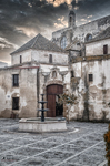 Plaza empedrada de Rota con iglesia blanca, portada histórica y cielo nublado.