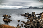 Costa rocosa del norte de España con mar tranquilo, piedras en la orilla e isla al fondo.