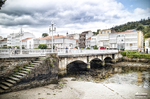 Pueblo marinero del norte de España con puente, casas junto al agua y cielo nublado.
