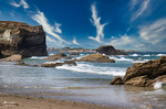 Playa de las Catedrales con acantilados, rocas sobre la arena y cielo azul atravesado por nubes.