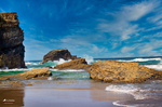 Rocas doradas sobre la arena húmeda en la Playa de las Catedrales de Ribadeo, con cielo azul y mar al fondo.