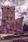 Torre medieval de Chaves rodeada de jardines, flores y cielo nublado.