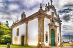 Pequeña capilla blanca en Chaves, con portada de piedra, jardín y cielo dramático.