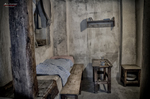 Celda austera de Terezín con cama estrecha, mesa pequeña y paredes deterioradas.