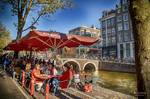 Terraza con sombrillas rojas junto a un canal de Ámsterdam en una escena luminosa y turística.