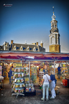 Puesto de recuerdos en Ámsterdam con turistas y torre histórica al fondo bajo cielo azul.