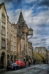 Calle escocesa con edificios antiguos, coches aparcados y torre de iglesia al fondo.