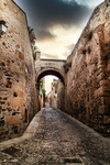 Calle empedrada entre muros de piedra con arco al fondo en Cáceres.