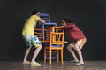Dos actores interpretan una escena teatral con sillas de colores sobre fondo negro.