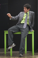Actor con traje gris y corbata verde sentado en una silla durante una escena teatral.