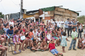 Niños y familias reunidos al aire libre durante una actividad teatral comunitaria de la compañía italiana Caravan.