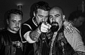 Tres hombres posan de noche, uno de ellos señalando a cámara, en ambiente motero.
