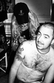 Mujer de pie junto a un hombre sentado en una escena íntima con tatuajes.