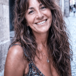Retrato callejero en primer plano de Alina, mujer madura de estilo bohemio, con cabello ondulado, sonrisa cálida y mirada directa en una calle antigua.