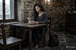 África permanece sentada ante una mesa de madera en una taberna rústica, con una taza entre las manos.